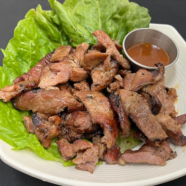 Grilled Thai BBQ pork slices on a bed of fresh lettuce with a side of dipping sauce.