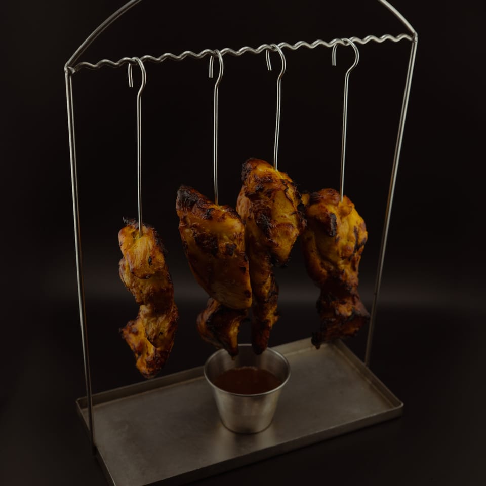 Grilled chicken pieces hanging from a metal stand over a dipping sauce cup.