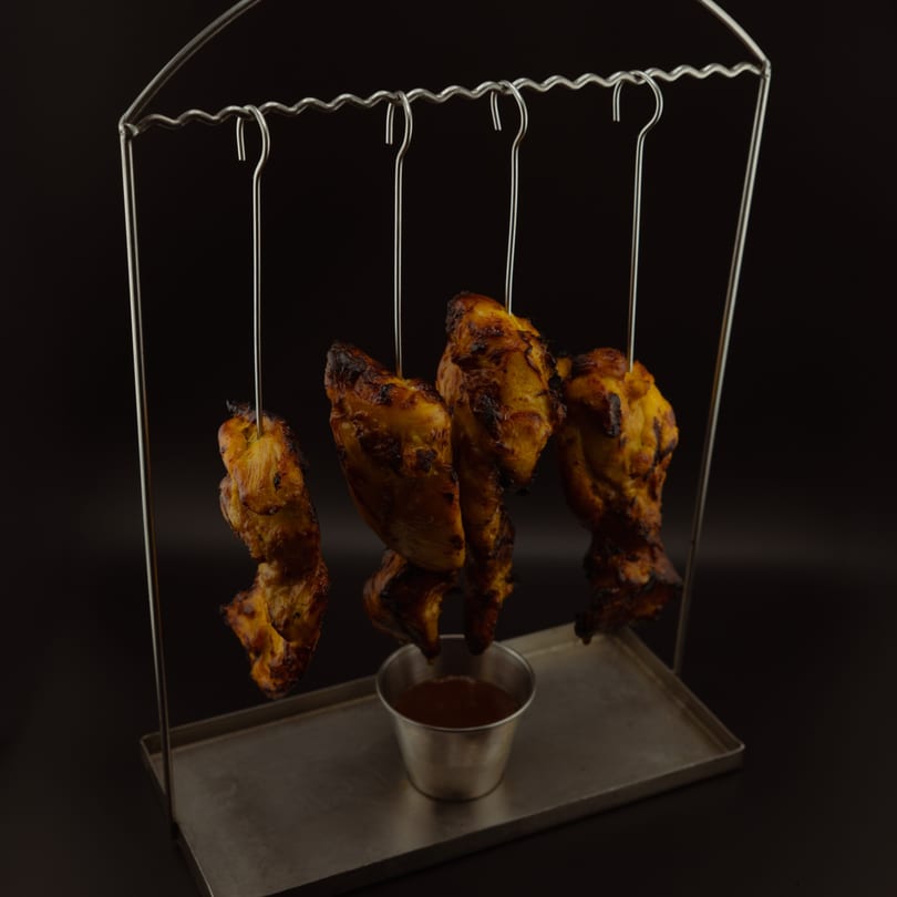 Grilled chicken pieces hanging from a metal stand over a dipping sauce cup.