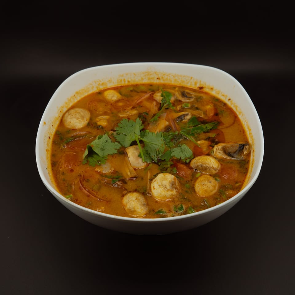 A white bowl of vibrant orange Thai Tom Yum soup with mushrooms and fresh cilantro.